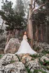 The bride with a bouquet of flowers stands on a rock in the mountain forest.