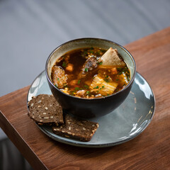 Fatty meat broth with ribs. Clay bowl of Hot soup and slices of black rye whole grain bread on blurred background.
