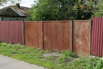one closed rusty iron gate with a closed door and part of a red metal fence on a rural street in green grass
