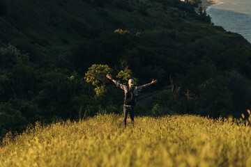 Young man with a backpack stands with raised hands on the top of the mountain, enjoying the view of the valley and forest area