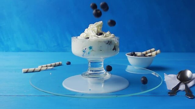 Blueberries fall into a glass ice cream cup that spins on a glass tray on a blue wooden table against a light blue background. Slow motion video with speed up, down effect.