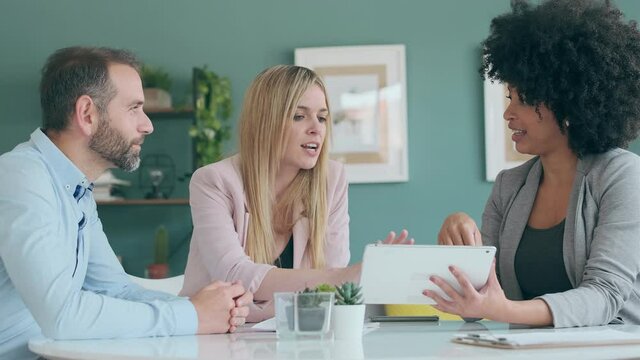 Video of beautiful real-estate agent showing house plans on electronic tablet while talking to the couple about buying the house in the office.