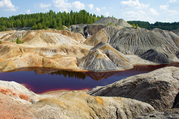 A dark red, acidic river against the backdrop of clay hills, a blue sky with clouds and a green forest. Hills, sky and forest are reflected in the surface of the water.