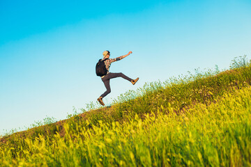 Happy young tourist with a backpack, happy jumping man with hands up against a blue sky background, climbs to the top of a hill or mountain