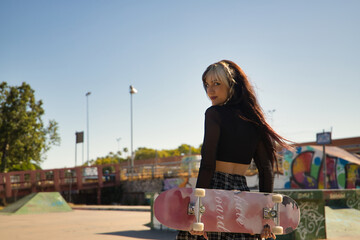Cute young girl with punk style and heterochromia holding a skateboard with her hands behind her back in a skateboard park. © Manuel