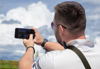 guy with his back turned makes a selfie on the phone. A bearded man in a white polo shirt with a smartphone in his hand takes a picture of himself against the backdrop of a cloudy sky. back view
