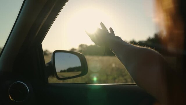 Girl with long hair is sitting in front seat of car, stretching her arm out window and catching glare of setting sun. Free woman travels by car catches wind with her hand from car window. Vacation