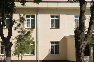 Wall of a beige building with closed windows on a sunny day, trees grow nearby