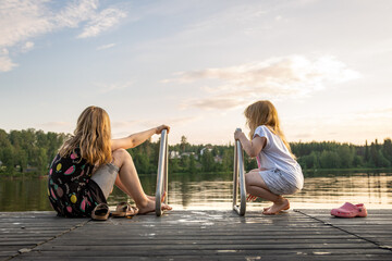Two young blonde girls on the edge of a dock pier on a lake during calm tranquil sunset