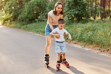 Full length portrait of woman and little son rollerblading together, mother teaching to roller skate his child, cute boy learning riding roller skates. - obrazy, fototapety, plakaty