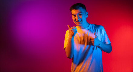 One Asian young man isolated on studio background in gradient pink purple neon light, colour filter. Concept of human emotions, facial expression.