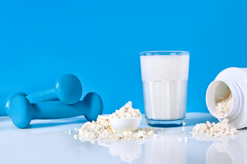 Glass of protein shake, protein powder in the spoon and blue dumbbells on a blue background. Copy space.