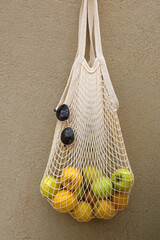 Mesh bag filled with lemons and green apples and retro sunglasses, hanging on a concrete wall.