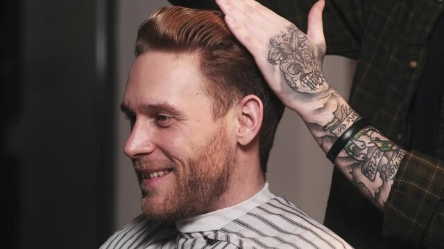 A cropped view of a tattooed hairdresser is doing hairstyle to his client while he looking to the mirror in a barber shop