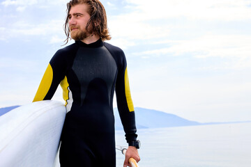 Young caucasian man with long hair and sportive body, walking with surf board on the beach, bearded athlete male is ready for surfing. Wearing stylish wetsuit, looking at side