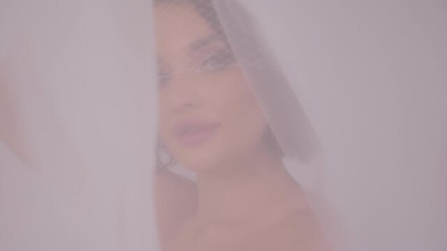 Close-up of the bride's face is covered with a veil, she looks at the camera.