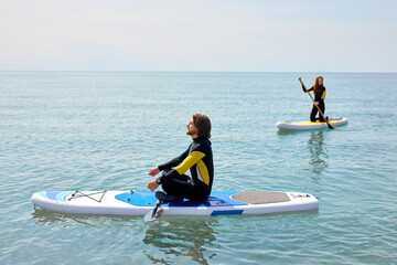 Paddleboard beach people on stand-up paddle boards have rest after surfing. Man and woman, caucasian couple enjoying watersport, wearing wetsuit. Extreme sport, leisure activity concept