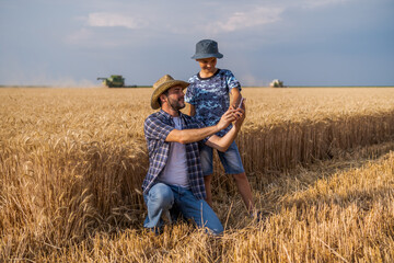 Naklejka na ściany i meble Farmers are standing in their wheat field while the harvesting is taking place. Father is teaching his son about agriculture.