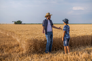 Fototapeta na wymiar Farmers are standing in their wheat field while the harvesting is taking place. Father is teaching his son about agriculture.