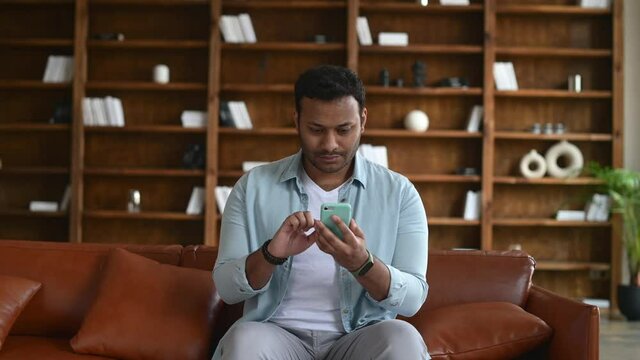 Indian young man sitting on the couch, holding and using smartphone, texting, chatting online, using new mobile app for dating or messaging, bookshelves on background