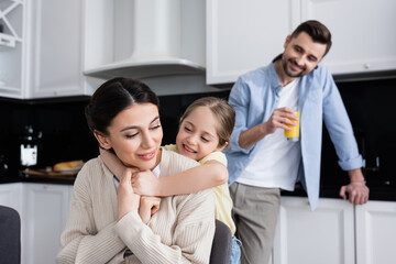 child embracing happy mom near dad standing with orange juice on blurred background