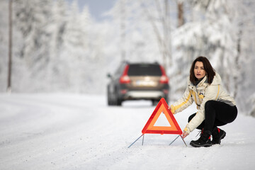 Pretty, young woman setting up a warning triangle and calling for assistance after her car broke down in the middle of nowhere on a freezing winter day