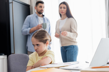 Obraz premium offended girl sitting with crossed arms near worried parents on blurred background