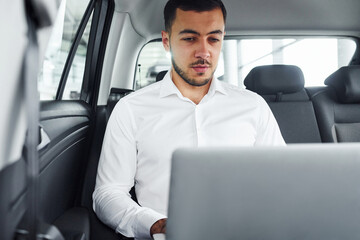 Works by using laptop. Young man in white shirt is sitting inside of a modern new automobile