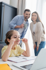 thoughtful girl doing homework near parents smiling on blurred background