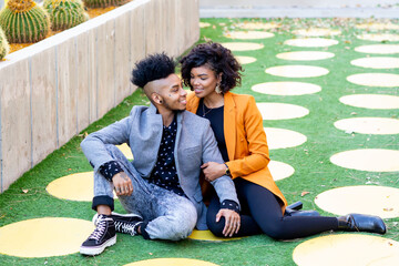 African American transgender couple sitting and smiling at each other on grass - obrazy, fototapety, plakaty