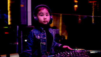 Plakat Asia little producer artist girl DJ in black headphones playing dance music and adjusting volume electronic sound mixer at the home party night
