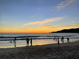 People relaxing on a sandy beach. Adults and children swim in the sea. Sunset, dark, twilight, evening time. Beautiful sky and orange glow of sun. Amazing clouds shape. Tourists on summer vacation.