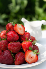 Blurred image of a plate with ripe strawberries on a white table with daylight in the nature background.