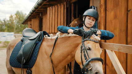 Female jockey fixing bridle of her light brown horse outside the stable.The girl wearing a safety...