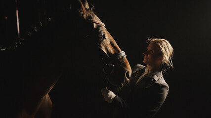 Female horse owner with her Dark bay Horse in a black background. Blond girl wearing gloves and dressed in a black coat fixing horse halter. Horse riding for leisure. 