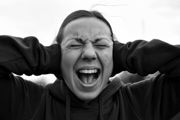 A young woman of 18-25 years old screams with her mouth open, covers her ears with her hands,...