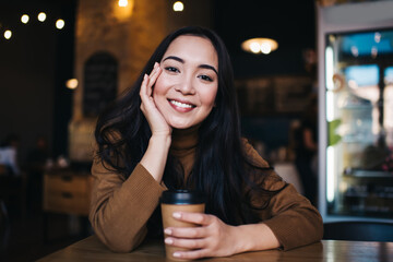 Portrait of prosperous female student enjoying coffee break in cafe interior sitting at table and posing, good looking hipster girl with caffeine beverage smiling at camera on leisure for recreate