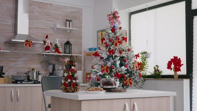Empty xmas decorated kitchen with nobody in it is ready for traditional christmas holidays. On table standing cookies dessert and cup of coffee beside christmas tree