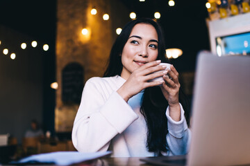 Contemplative freelance copywriter pondering on ideas for web publication sitting at cafeteria table with modern laptop computer and dreaming, hipster girl enjoying distance job in coffee shop