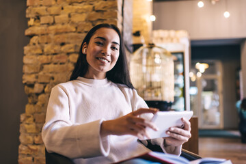 Positive young lady holding tablet in hands while sitting at cafe and looking at camera