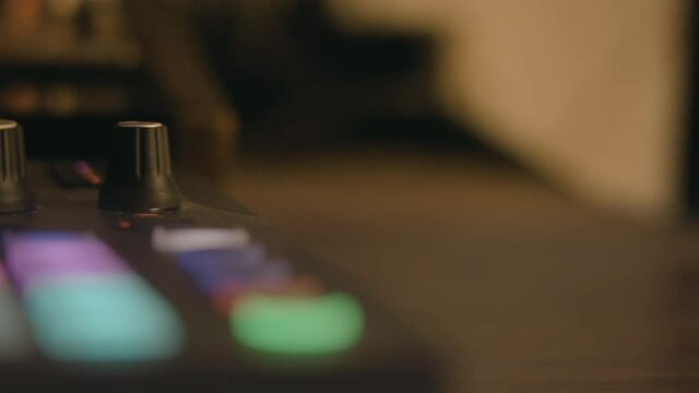 Smooth slow-motion panning through the knobs of a synthesizer, the light blue and pink buttons of the sequencer are out of focus. One hand touches knobs and buttons