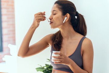 Sporty young woman eating iogurt while standing in the living room at home.