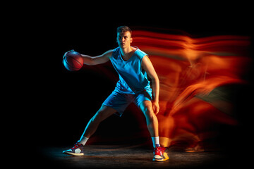 Fototapeta na wymiar Young caucasian male basketball player playing basketball isolated over dark studio background in mixed light. Concept of healthy lifestyle, professional sport, hobby.
