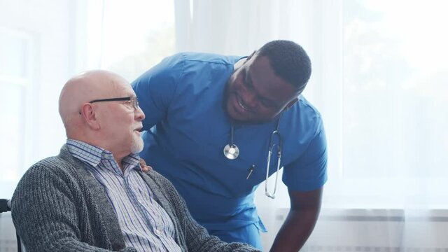 African-American caregiver and old disabled man in a wheelchair. Professional nurse and handicapped patient in a nursing home. Assistance, rehabilitation and health care.