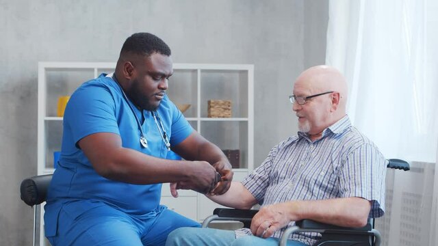 African-American caregiver is measuring disabled old man blood pressure. Professional nurse and handicapped patient in a nursing home. Assistance, rehabilitation and health care.