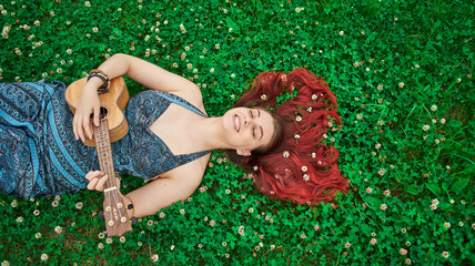 Top view portrait of woman playing ukulele lies in a meadow in summer time, red hairs are scattered...
