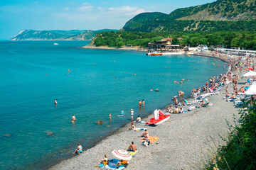 Beautiful coast of the Black Sea with a crowd of tourists on a background of green hills. The beach with many tourists resting in the sun with hot weather.
