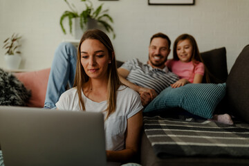 Fototapeta premium Mom freelancer is working at home, sitting on the floor while dad and daughter having fun on sofa.