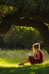 sunny portrait of a beautiful girl sitting on green glade under an arch of tree branches with book,...