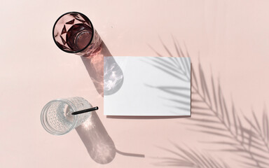 Summer tropical mockup still life flat lay. Empty blank invitation card, cocktail glasses on pink background. Palm leaves shadow. Mock minimal concept for wedding, vacation or relax. Reflection.
